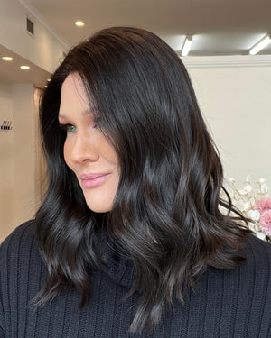 Person with long dark hair standing indoors, looking at a mirror.