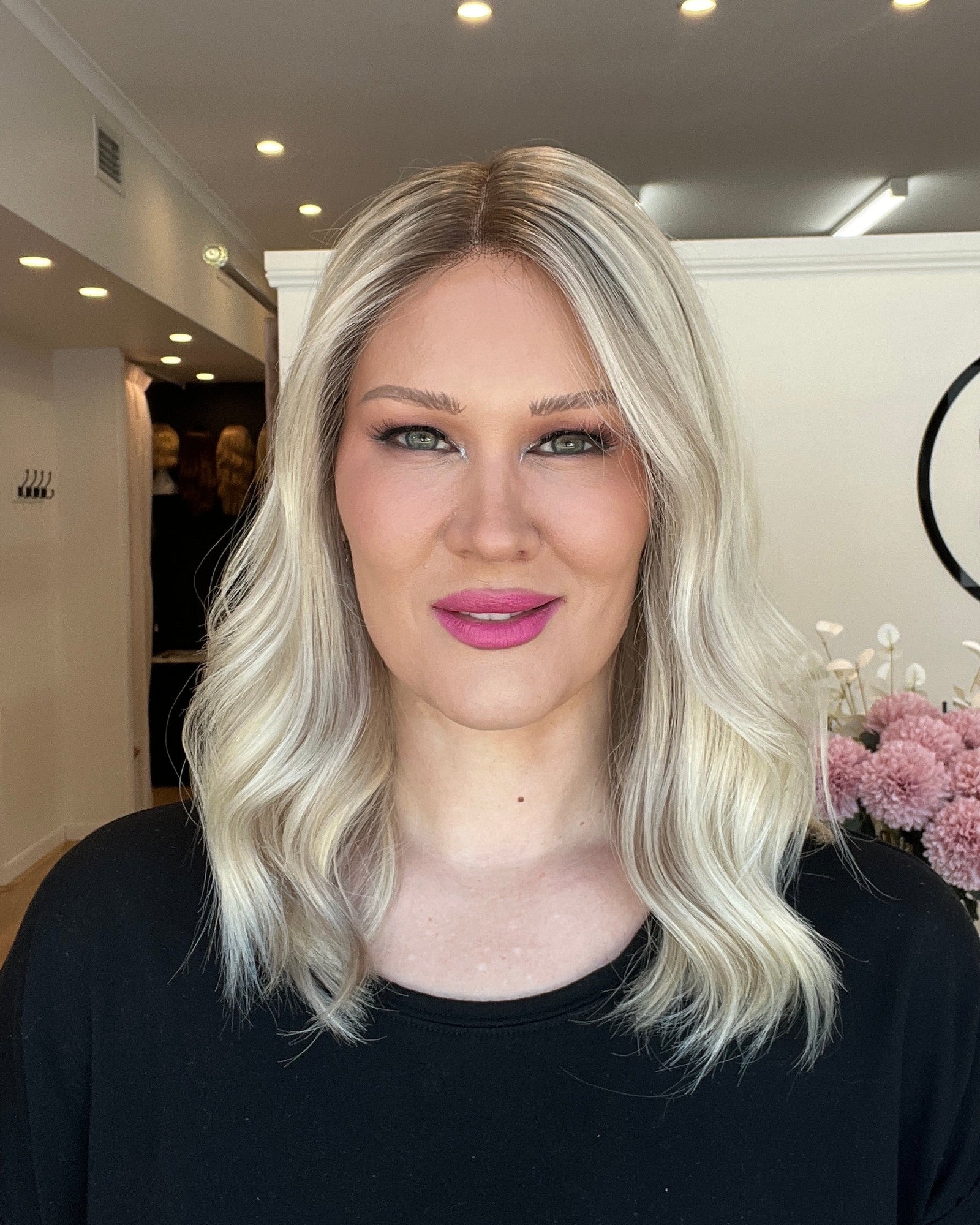 Person with blonde hair and pink lipstick standing indoors, wearing a black top.