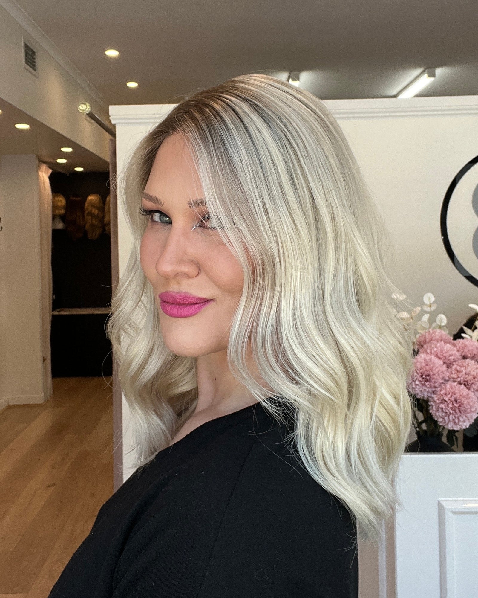 Person with blonde hair and pink lipstick standing indoors, wearing a black top.