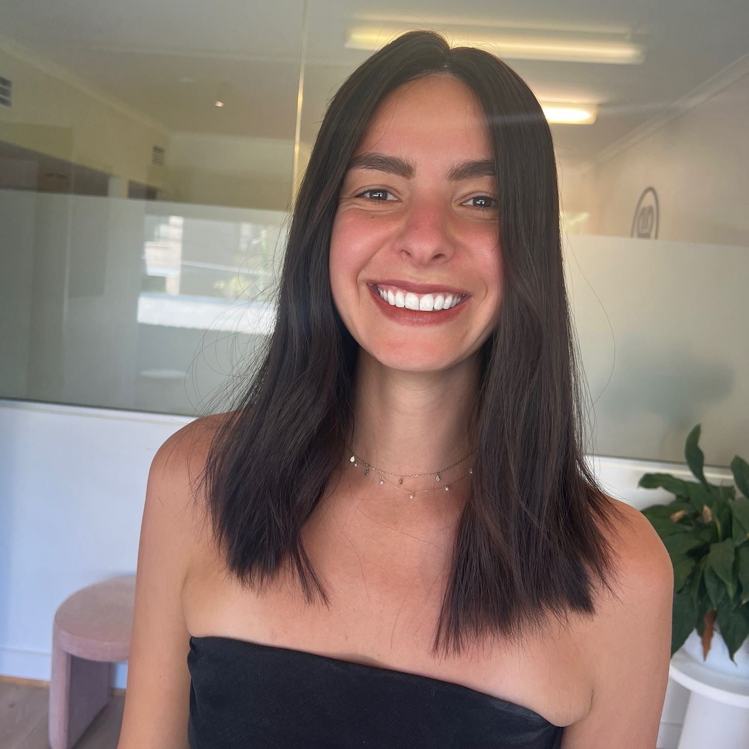 Woman with long dark hair wig smiling in a room with a glass wall and plant.