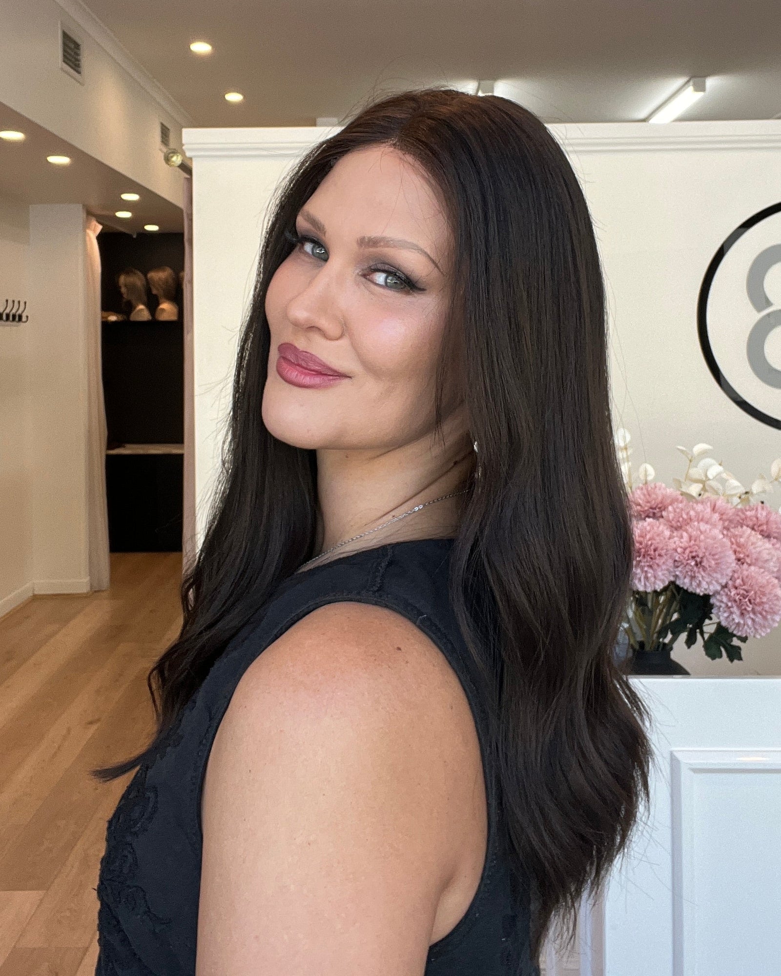 Woman with long dark hair and pink lipstick standing indoors, looking at the camera.