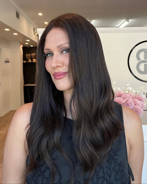 Woman with long dark hair and pink lipstick standing indoors, looking at the camera.