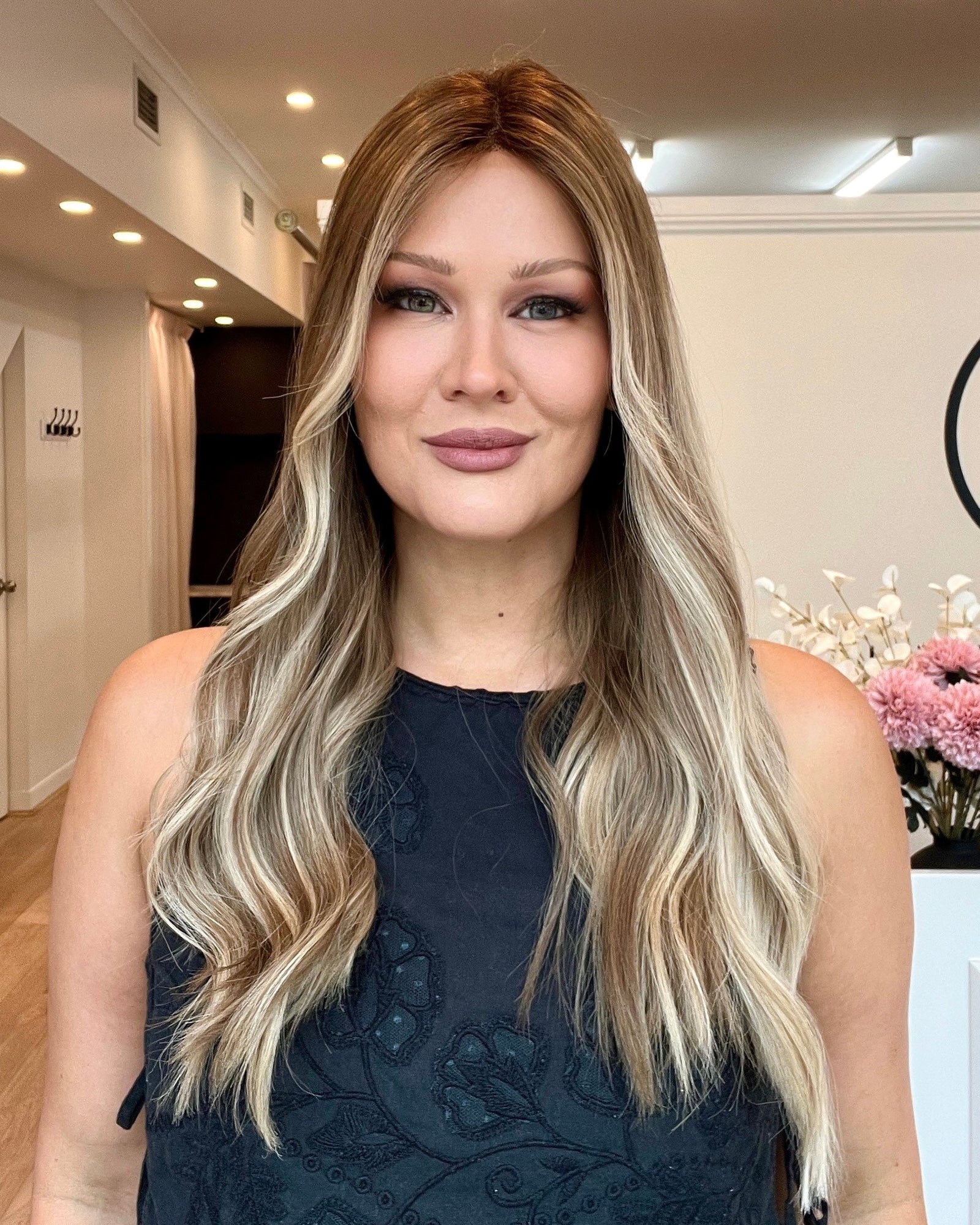 A woman wearing a black sleeveless dress with a floral pattern, featuring long wavy blonde hair with highlights.