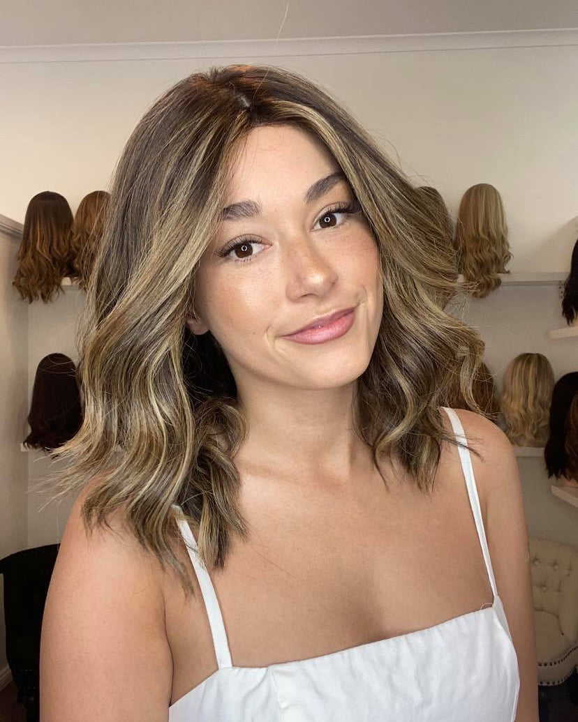 A woman wearing a light brown wig with a silk top and lace front, reaching up to her shoulders.